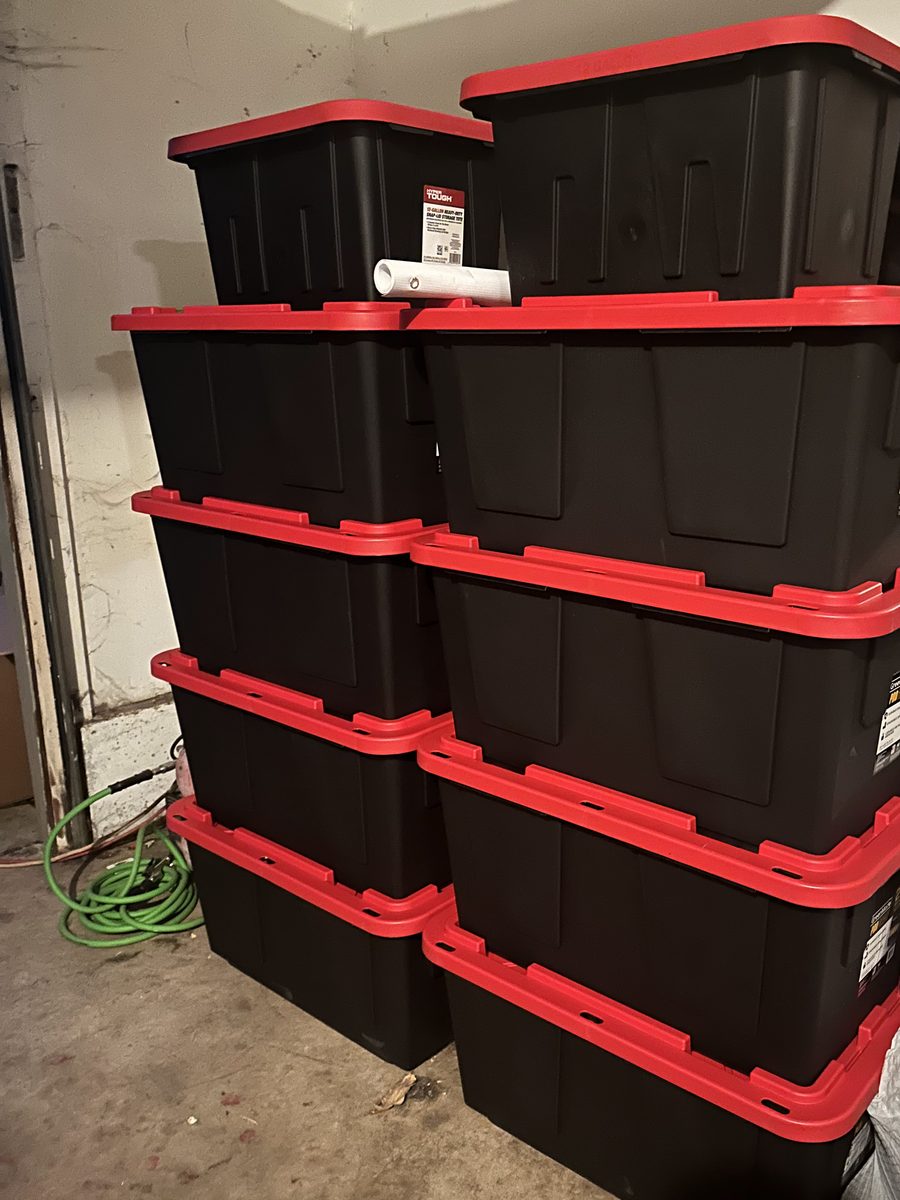 Ten black storage totes with red lids, stacked in two columns in a corner of the garage.